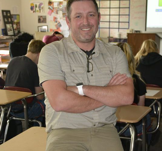 Jim Reed poses on a desk in his classroom with students working in the background
