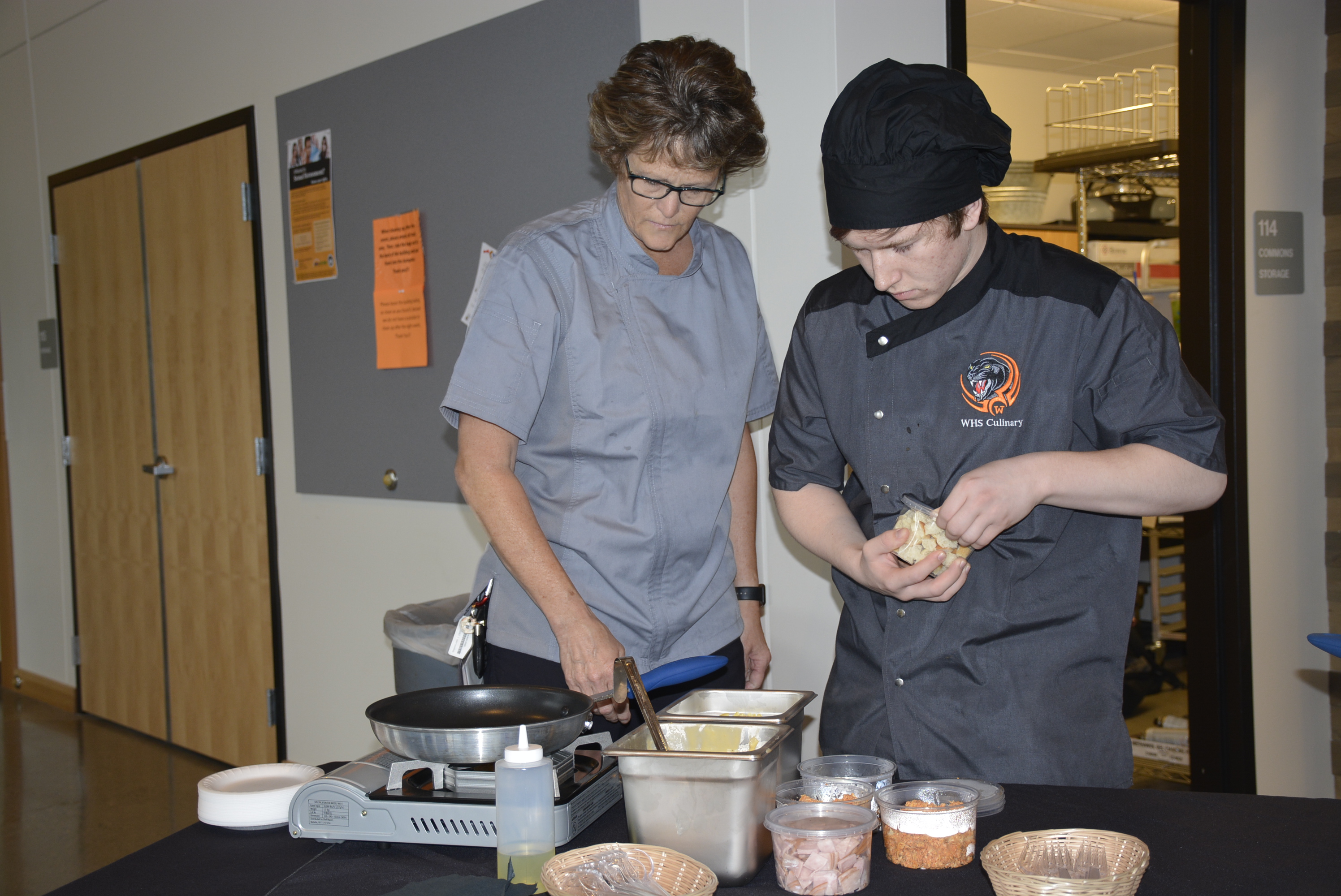 Brenda and a student in front of a table where student is preparing food as part of their culinary final