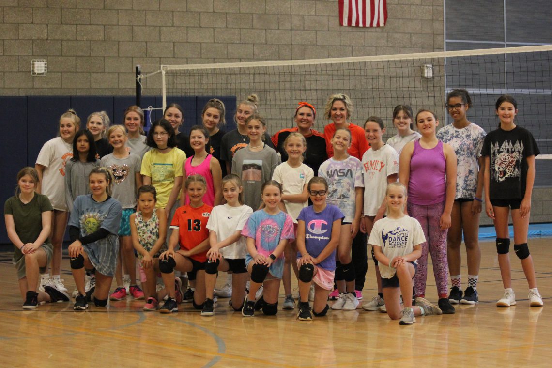 Twenty-six students and 2 coaches form three rows to pose for a group photo. Students are all girls ranging from middle to high school age. Girls in the front row kneel on one knee. Two adult female coaches are in the back row in the center of photo. Group is indoors on light brown laminate gym flooring with a volleyball net set up behind them.