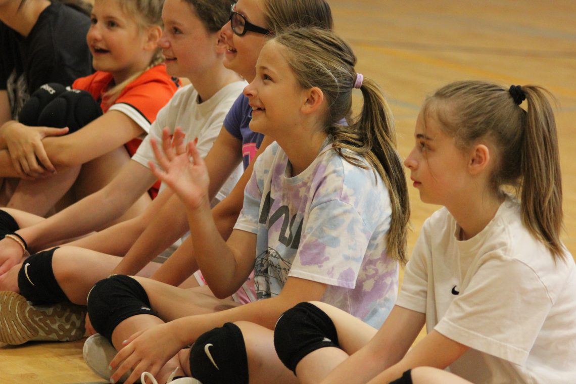 Five girls sit on laminate gym floor facing left. They are aligned diagonally, in a row beginning in the upper left corner and ending in the lower right corner of image. They are wearing t-shirts, shorts and knee pads. They all smile.