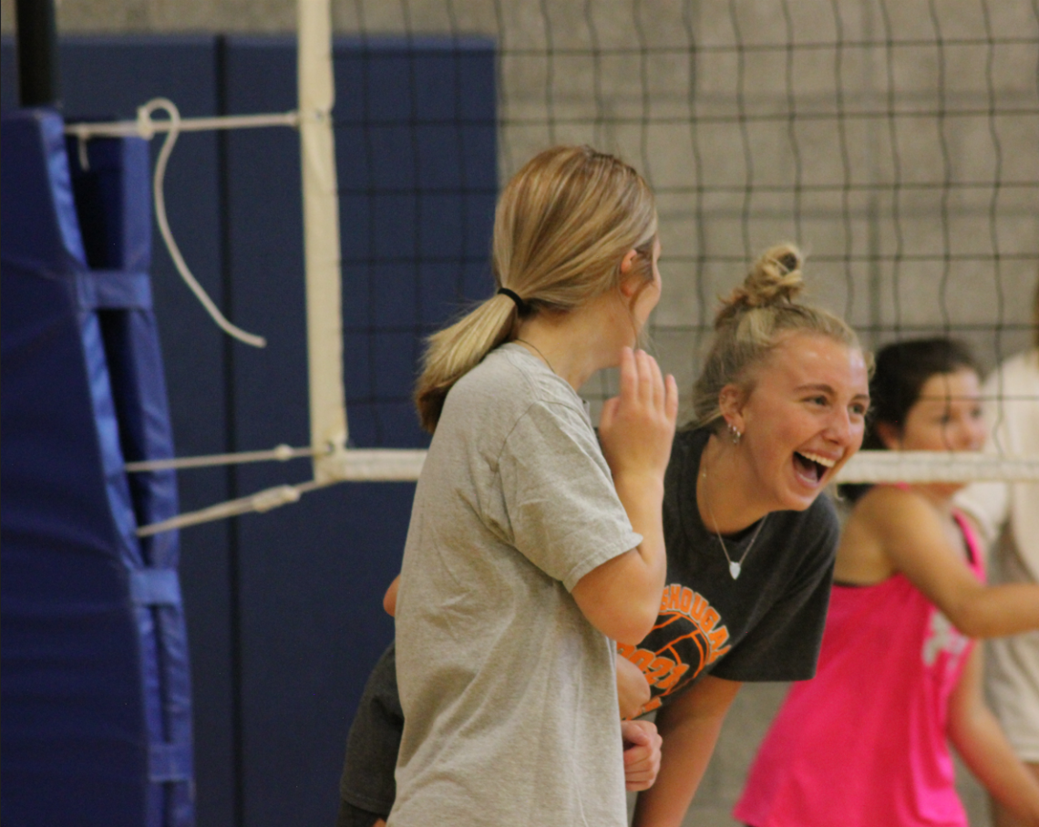 Two high school girls stand side by side in the indoor gym. The girl closest to the camera faces away from the camera to the second girl. The second girl leans forward with laughter. In the background is a volleyball net and a younger girl running.