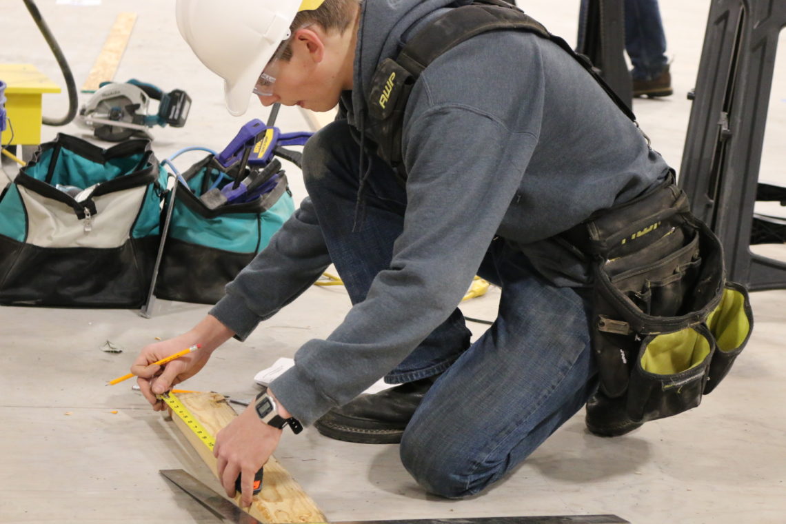 High School student kneels to measure a board of wood, wearing a hard hat.