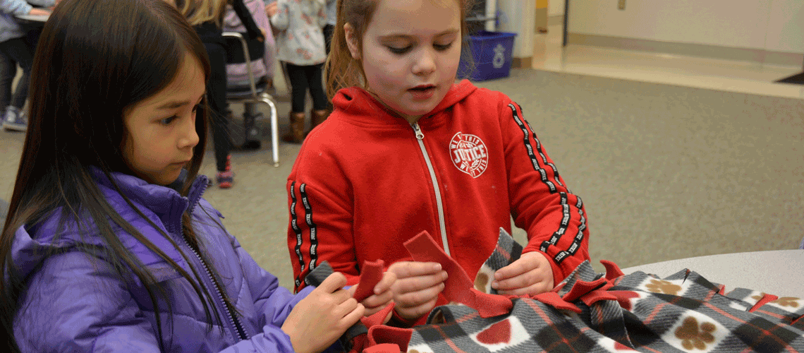 Two students tie knots in the edge of a blanket they are making, seated at a table