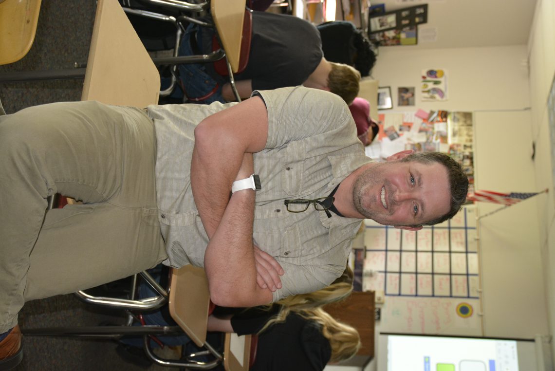 Jim Reed poses on a desk in his classroom with students working in the background