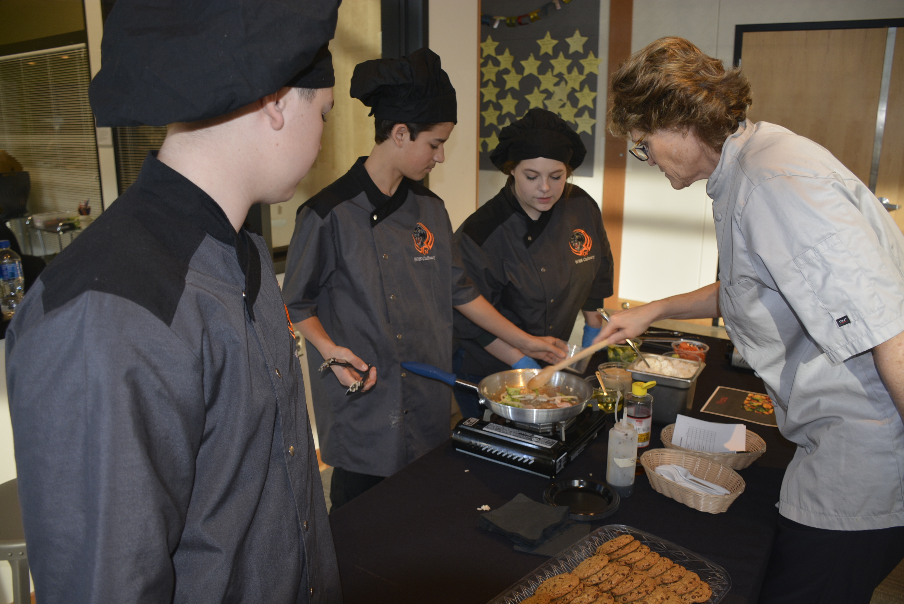 Brenda and three students in front of a table where student is preparing food as part of their culinary final