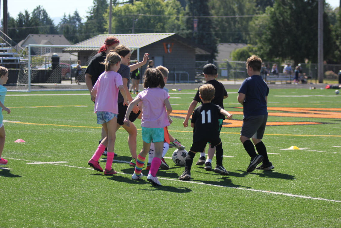 Six elementary school aged athletes run after a soccer ball. They are positioned in a group with the soccer ball at the center. Two high school coaches are on the left side of the group.