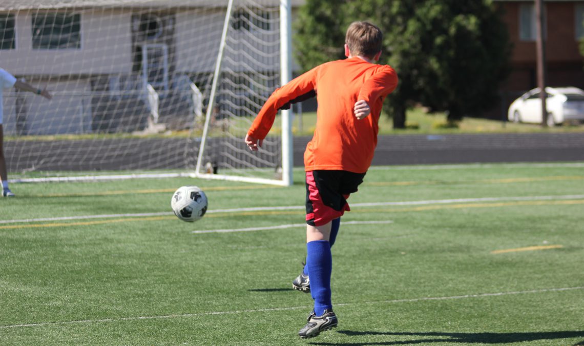 Moment is captured just after middle school boy kicks soccer ball toward the goal. Boy is outdoors. The ball flies about 20 feet ahead of him. Boy faces away from camera. Ball is to left of boy. Soccer goal can be seen in the background.