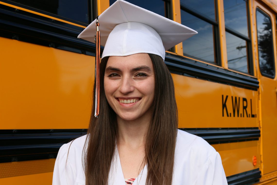 Young woman with long brown hair and white graduation cap and gown faces camera. Woman is shown from shoulders up. In background, yellow bus fills frame.
