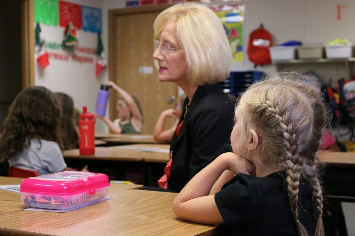 Superintendent Dr. Mary Templeton sits at a desk with blonde 1st grade girl. Girl in foreground has two french braids going down the back of her head and a black shirt. Two students in background sit at desks and look attentive.