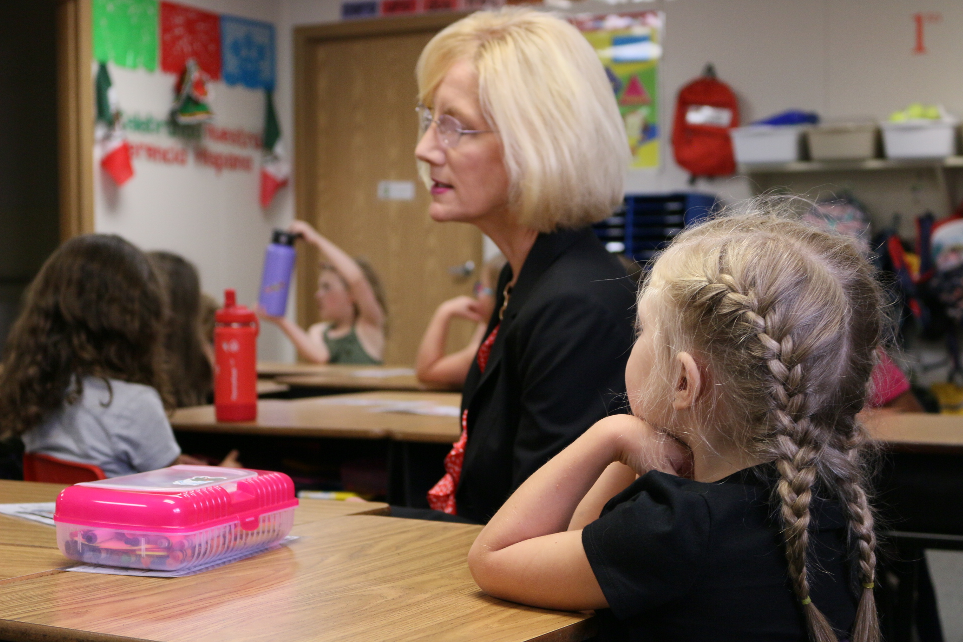 Superintendent Dr. Mary Templeton sits at a desk with blonde 1st grade girl. Girl in foreground has two french braids going down the back of her head and a black shirt. Two students in background sit at desks and look attentive.