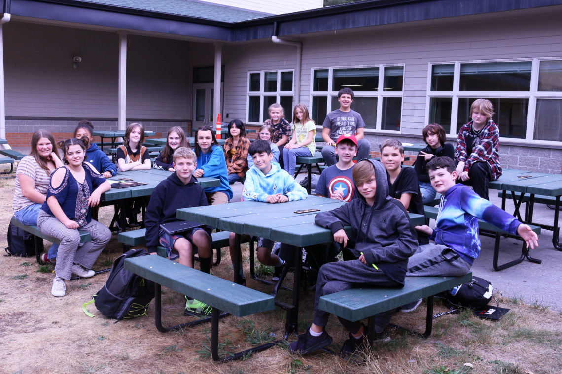 Group of about 30 students sit at picnic tables outdoors and face toward camera.