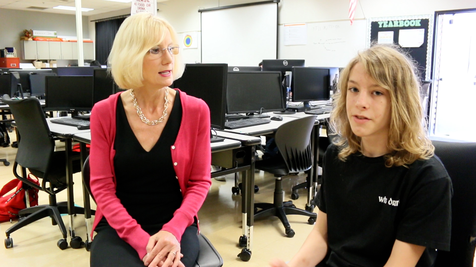 Superintendent Mary Templeton sits with high school student Raphael indoors with computer monitors on desks in the background.
