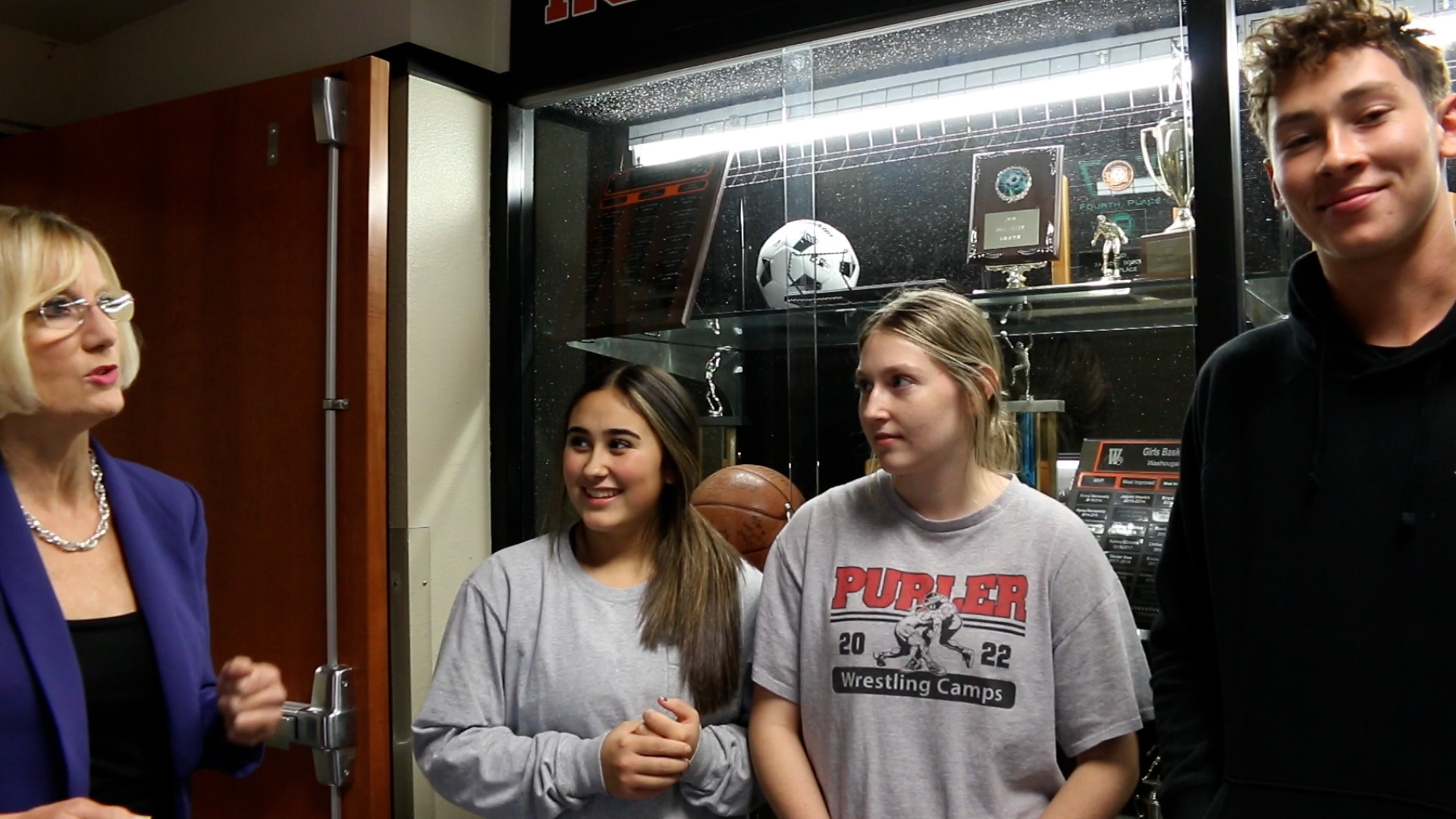 Three students stand with the superintendent indoors in front of trophy case.