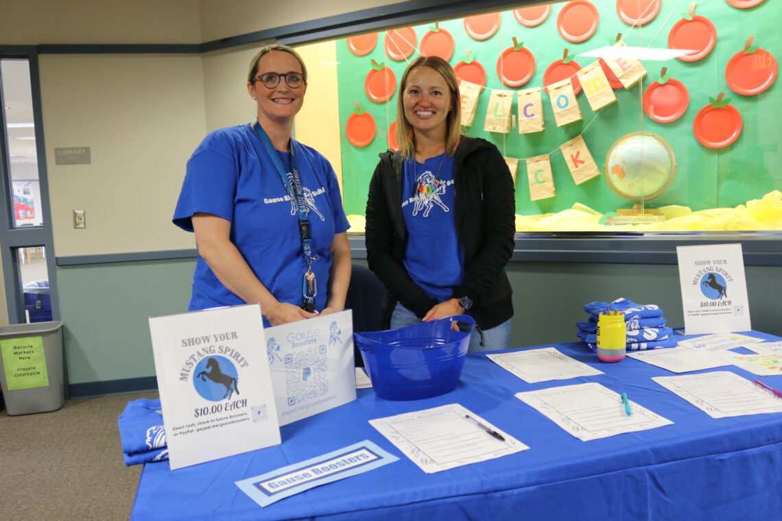Two women in royal blue school shirts smile and stand behind a table with volunteer materials and a blue table cloth on it.