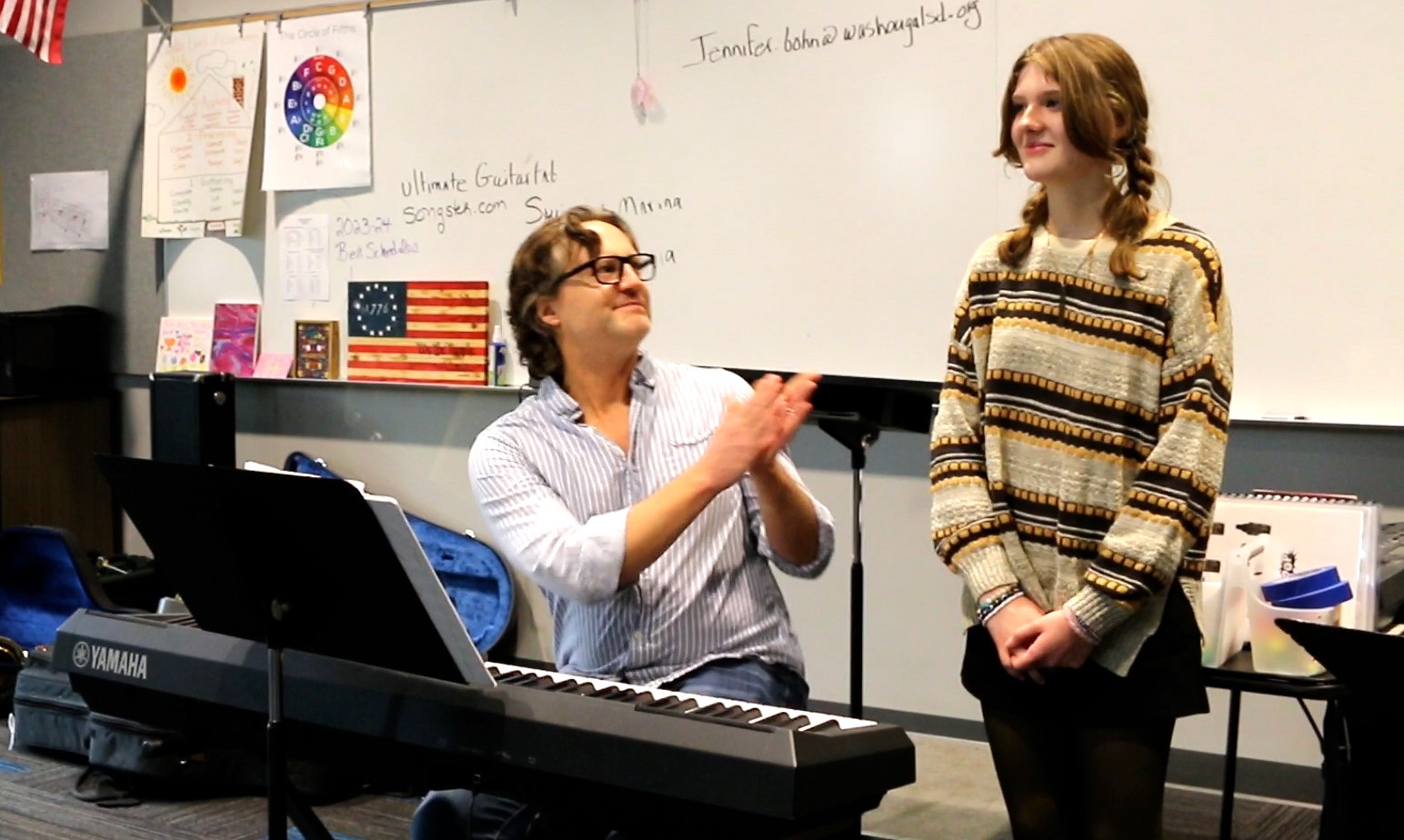 Adult sitting at piano claps while smiling at standing student next to him.